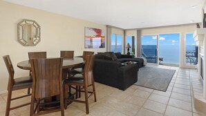 A spacious living area with a dining table and chairs, a black sofa, and a large window offering a view of the ocean.