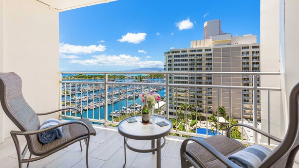 A balcony with two chairs, a small table with a bottle and flowers, and a view of a marina with boats and a hotel in the background.