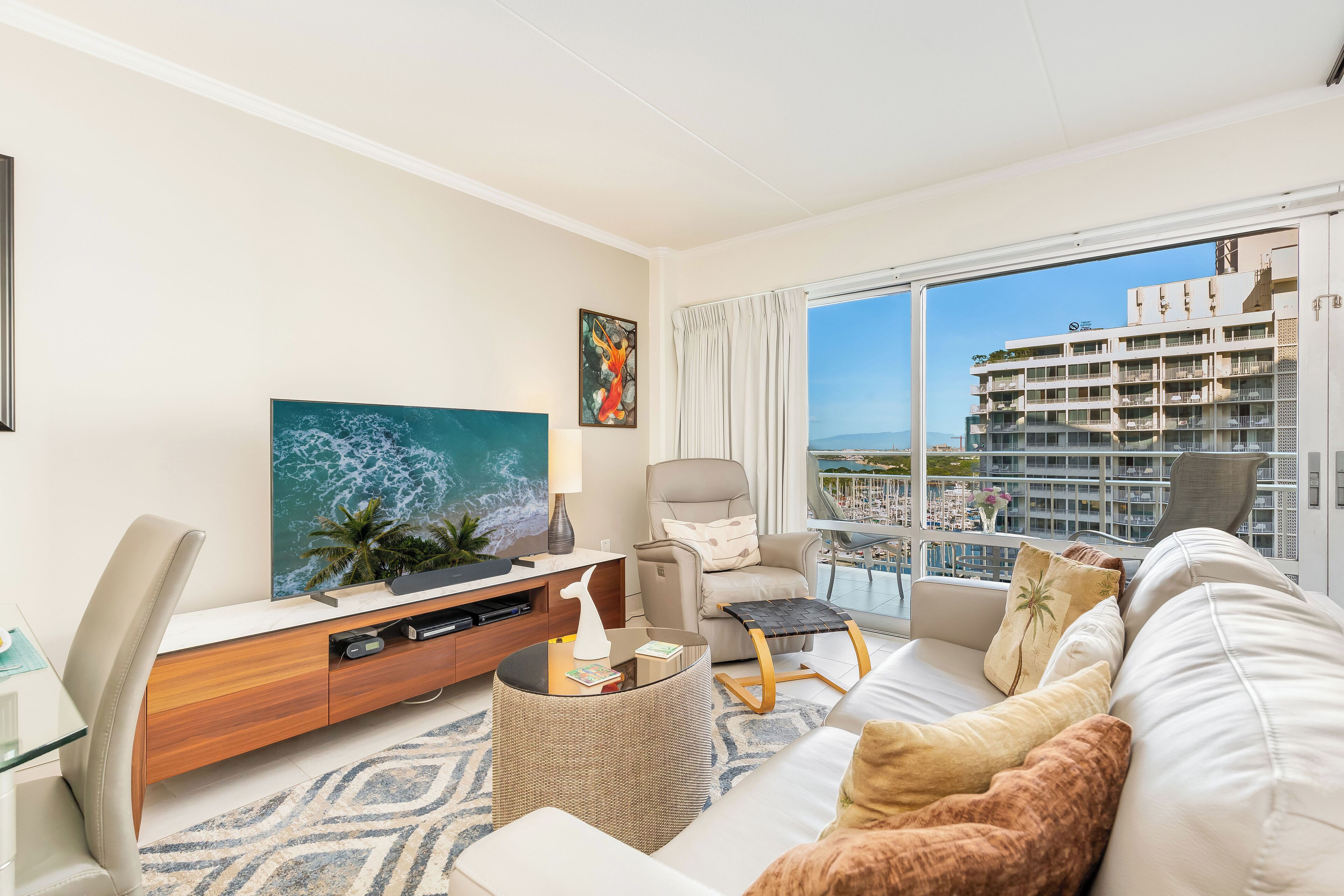A modern living room with a large flat-screen TV, a comfortable sofa, a chair, and a balcony with a view of a high-rise building.
