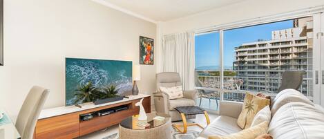 A modern living room with a large flat-screen TV, a comfortable sofa, a chair, and a balcony with a view of a high-rise building.