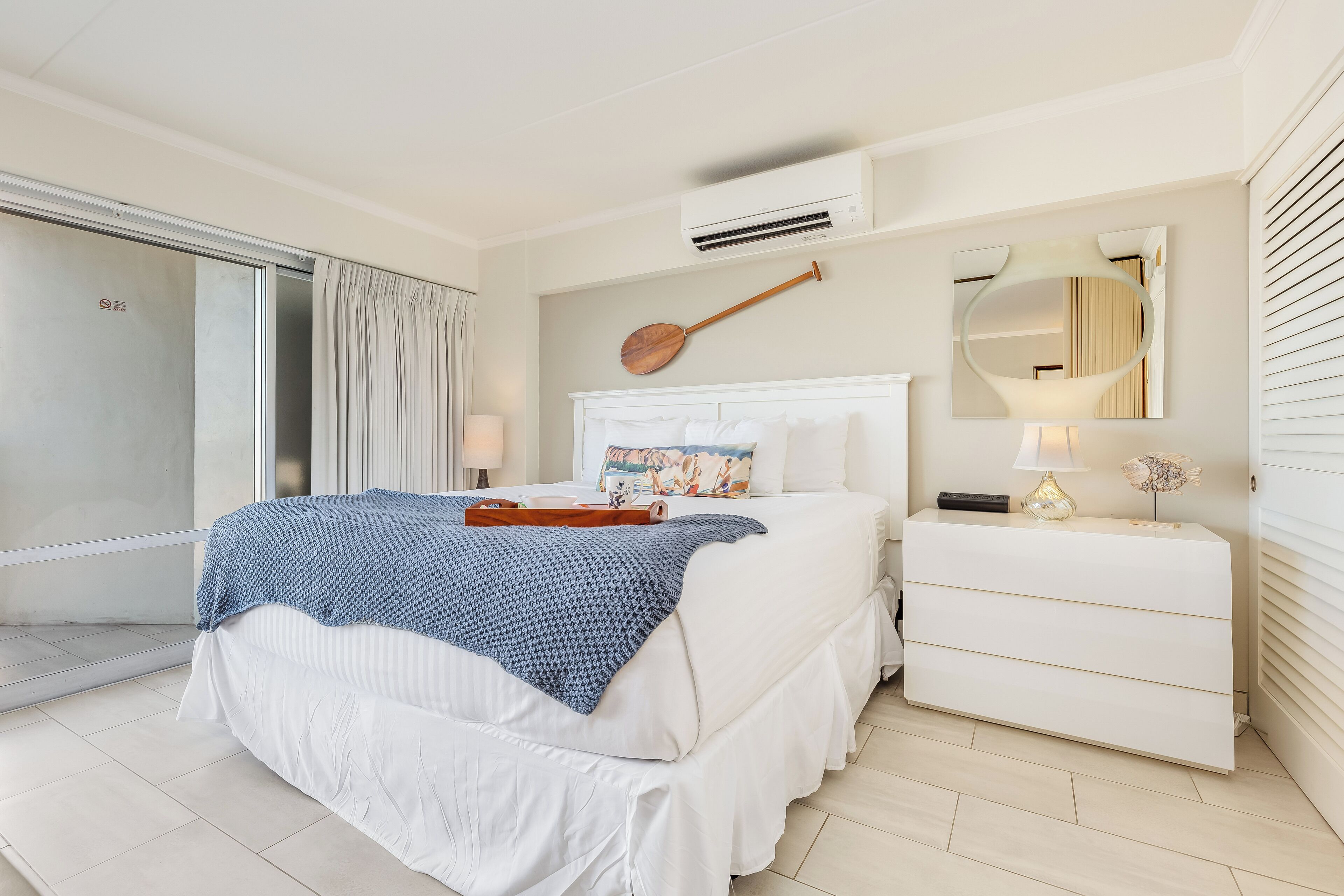 A hotel room with a bed, a bedside table, a mirror, and an air conditioning unit.