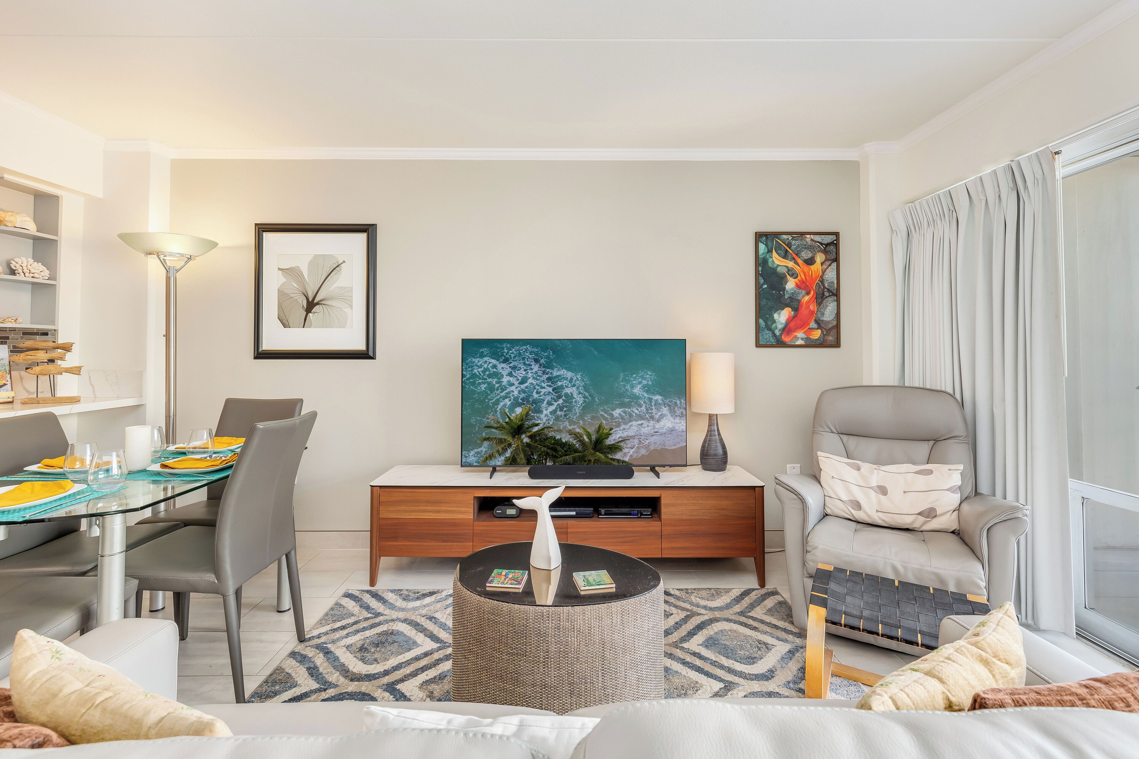 A modern living room with a glass dining table, comfortable seating, a flat-screen TV, and artwork on the walls.