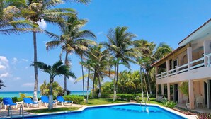 A swimming pool with blue water, surrounded by palm trees and a building with a balcony. - Stunning Caribbean views from the wrap around deck, at this beachfront condo! (South Akumal Beach)