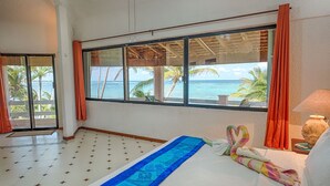 A room with a large window offering a view of the ocean and palm trees. There is a bed with a blue bedspread and a decorative pillow. - Stunning Caribbean views from the wrap around deck, at this beachfront condo! (South Akumal Beach)
