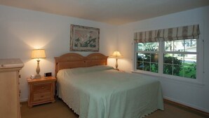 Een slaapkamer met een bed, twee nachtkastjes met lampen, een nachtkastje, een schilderij aan de muur, en een raam met uitzicht op groen.
