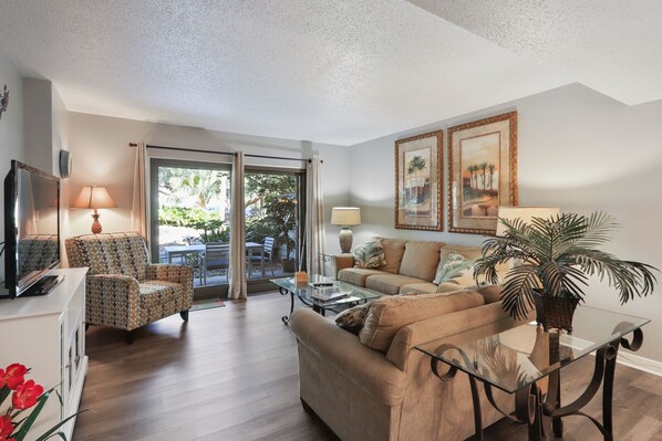 A living room with a sofa, armchair, TV, and a glass coffee table. There are two lamps and a potted plant.