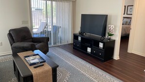 A living room with a leather sofa, a black entertainment unit with a flat-screen TV, a patterned rug, and a sliding glass door leading to a patio.