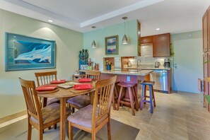 Un'area pranzo con sedie di legno e un tavolo apparecchiato per quattro, una cucina con mobili in legno e un piano di lavoro, e un quadro appeso al muro.
