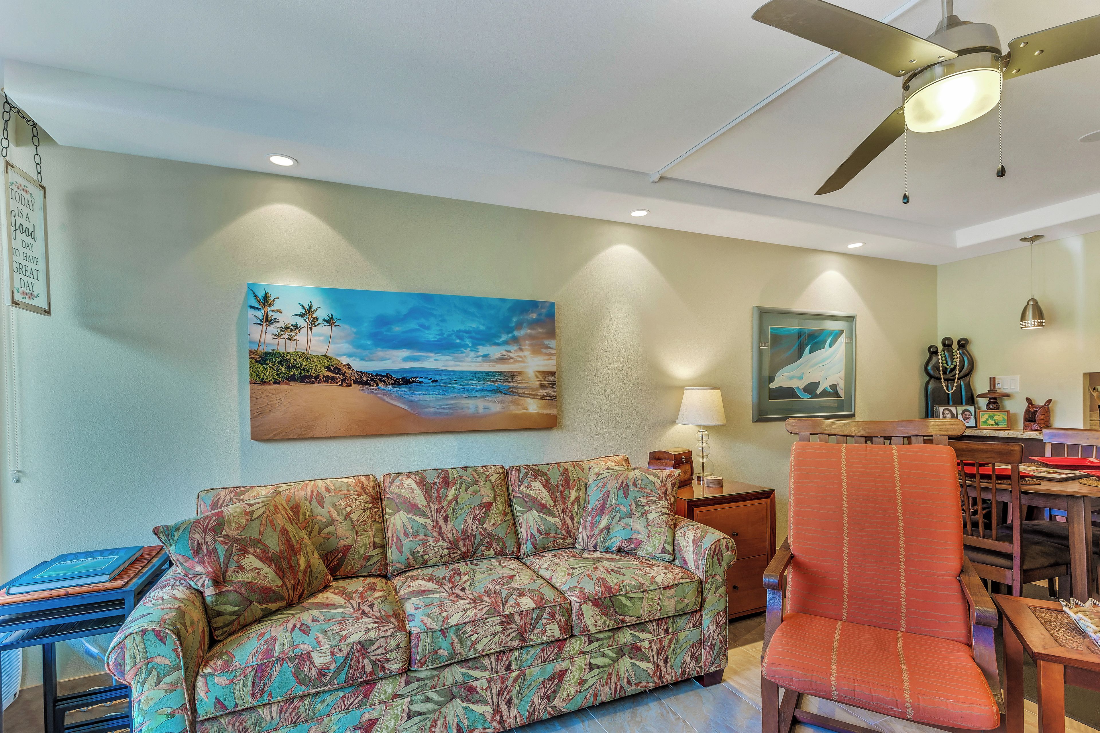 Un salon avec un canapé, un fauteuil, un ventilateur de plafond et un tableau représentant une scène de plage.