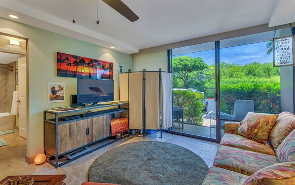 A modern living room with a flat-screen TV, a sofa, a coffee table, and a large window offering a view of greenery.