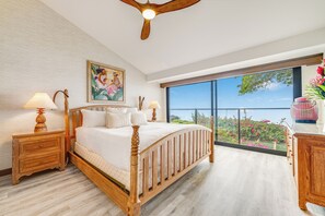 A bedroom with a wooden bed, two bedside tables, a large window with a view of greenery, and a ceiling fan.