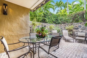 Une terrasse extérieure dotée d’une table ronde en verre, entourée de chaises en métal, d’un mur en pierre et d’une végétation tropicale luxuriante.