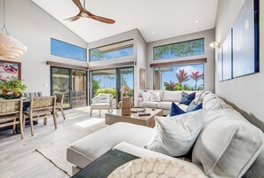 A modern living room with a large sofa, a coffee table, and a dining area with a bench and chairs. There are large windows with views of greenery and a ceiling fan.