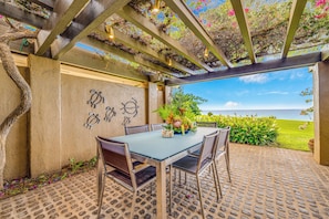 Un espace extérieur abrité, aménagé pour les repas, avec une table et des chaises, une vue sur l’océan et une pergola couverte de plantes grimpantes.