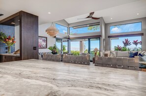 Amplia sala de estar con piso de mármol, asientos integrados, ventanas grandes, ventiladores de techo y plantas en macetas.