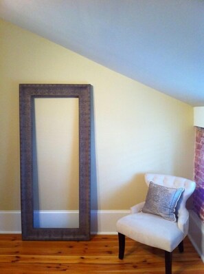 A room with a large mirror, a chair with a cushion, wooden flooring, and a sloped ceiling.