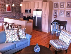 A kitchen with a refrigerator, microwave, and island with stools. There is a blue sofa and patterned armchair in the living area. - "Josephine" Historic Arts District Condo, Walk to Smoothie King, Superdome & Morial Convention Center, Ports and view of WWII Museum, its that close (New Orleans)