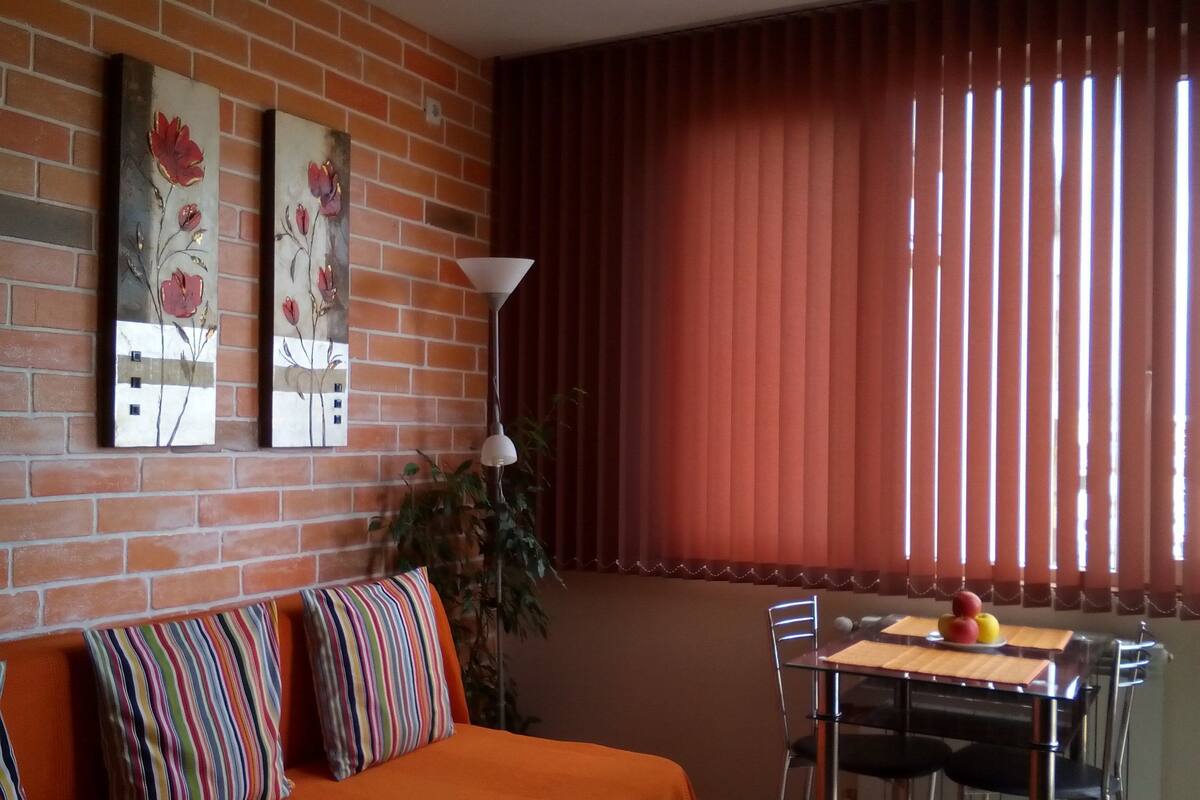 A room with a brick wall, a sofa with colorful pillows, a dining table with chairs, and a window with vertical blinds.