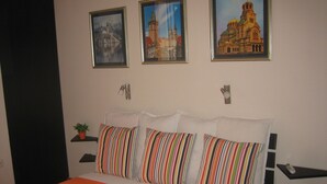 Une chambre d’hôtel avec un lit, des coussins à rayures, une plante en pot et trois tableaux encadrés accrochés au mur.