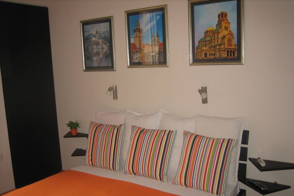 A hotel room with a bed, striped pillows, a potted plant, and three framed pictures on the wall.