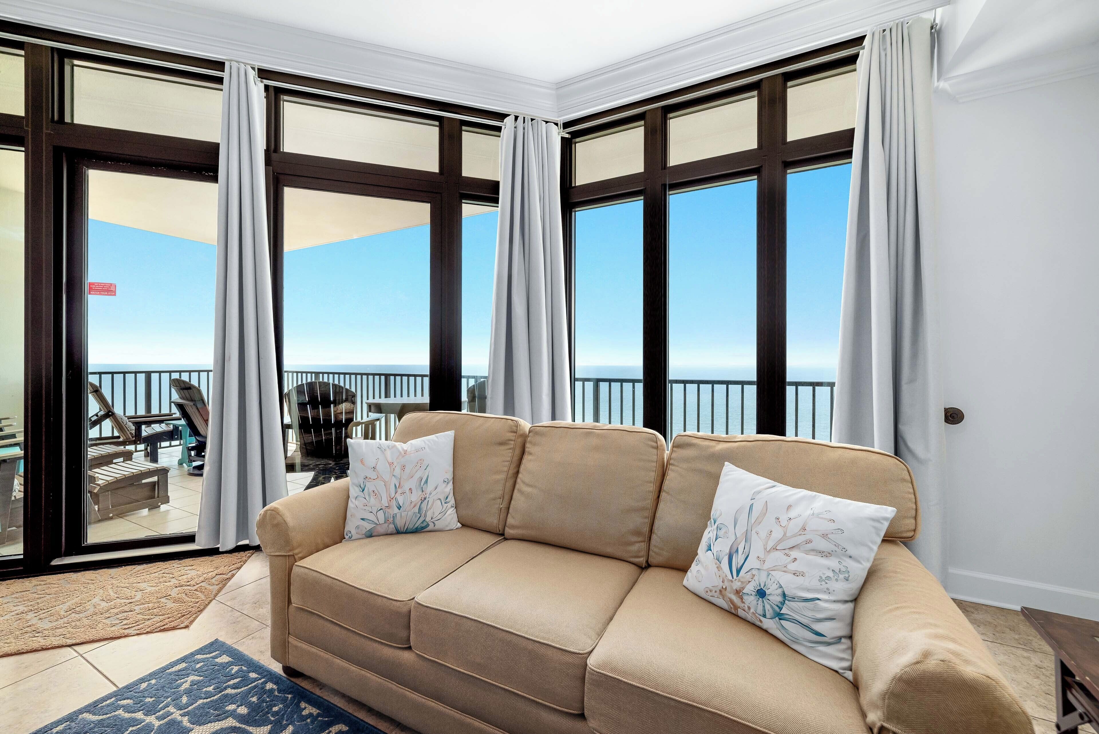 Ein Zimmer mit einem beige farbenen Sofa, zwei dekorativen Kissen, einem blauen Teppich mit Muster und Meerblick durch große Fenster mit Vorhängen.