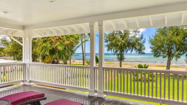 Un espacio al aire libre techado con vista a la playa, que incluye una barandilla blanca y una zona de estar con sillones acolchados.