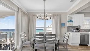 A dining area with a view of the ocean, a chandelier, and a kitchen with white cabinets.