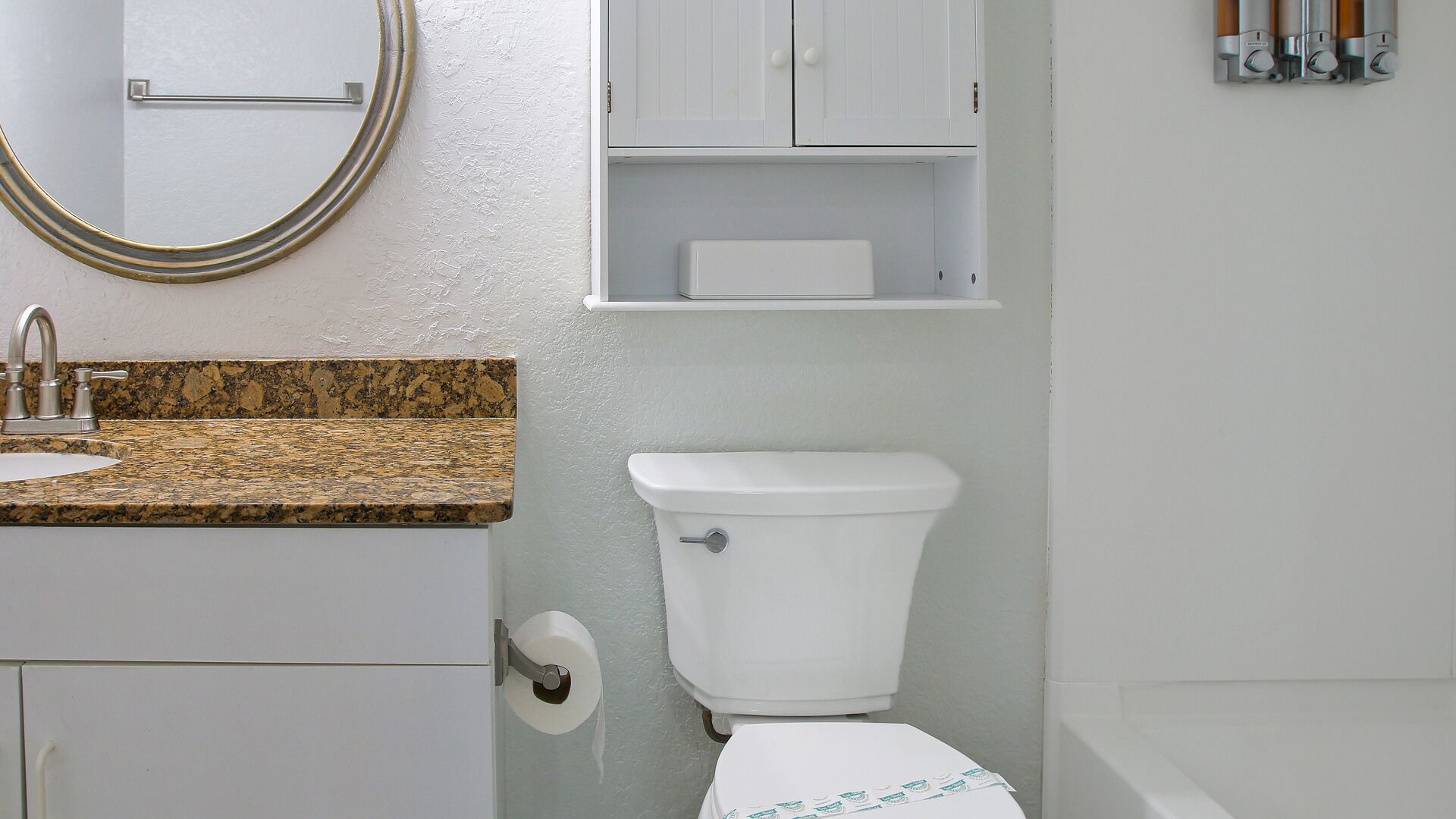 Un baño con lavamanos de encimera de granito, inodoro y un mueble blanco sobre el inodoro.
