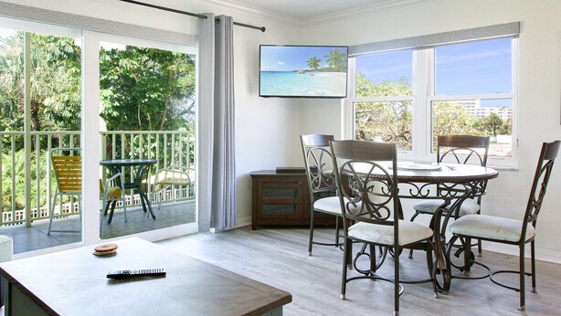 Un comedor con mesa y sillas, un televisor en la pared, y vistas a la vegetación y una playa a través de amplios ventanales.