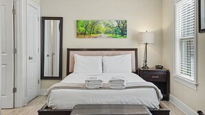 A bedroom with a bed, a bedside table, a lamp, a window with blinds, and a painting on the wall. - Modern Suite for the Traveling Professional by Lucky Savannah (Savannah)