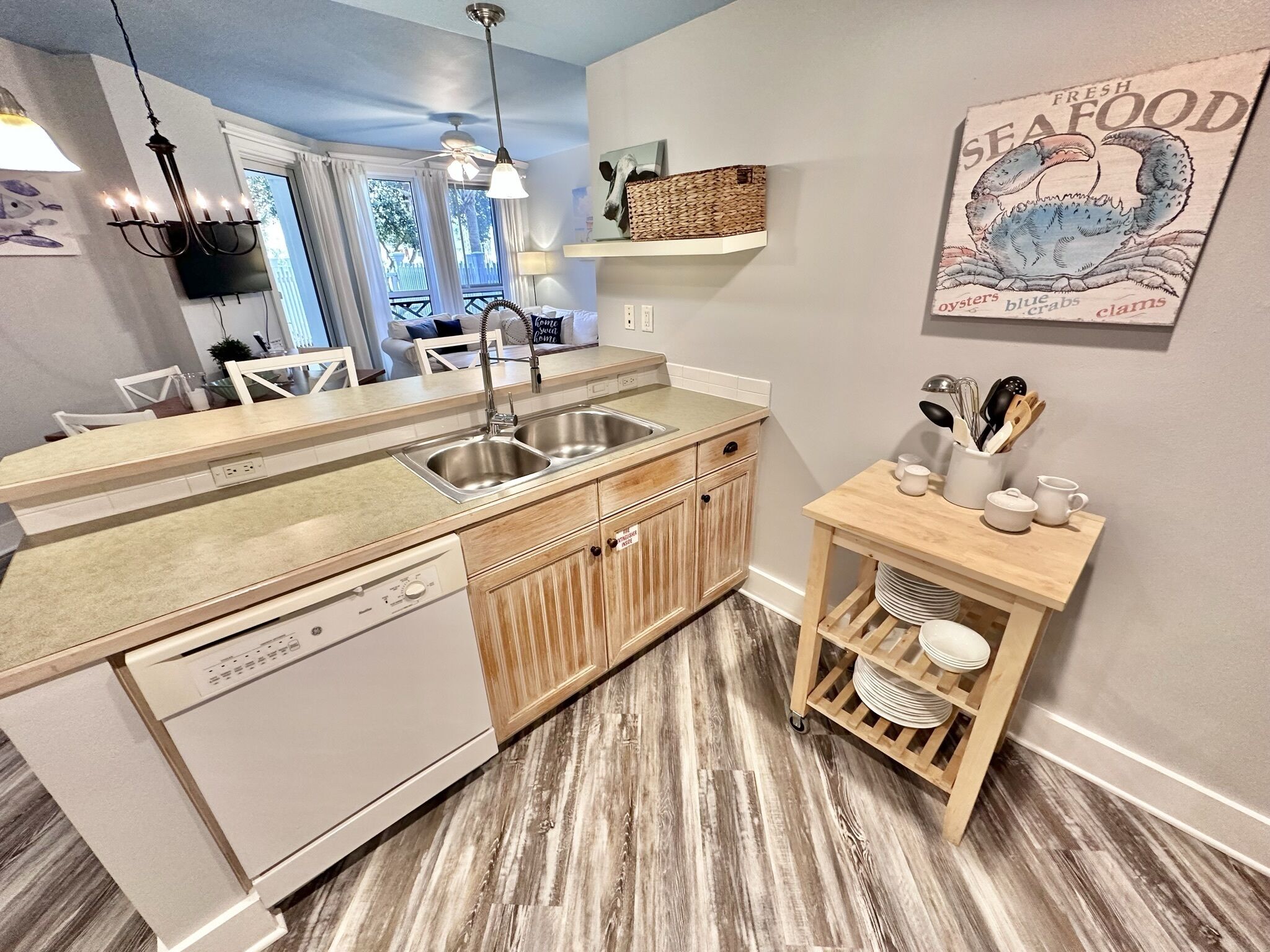 A modern kitchen with a dishwasher, double sink, wooden cabinets, and a small wooden table with kitchen utensils.