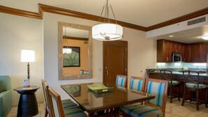 A dining area with a wooden table, striped chairs, a chandelier, and a kitchen in the background. - Maui Resort Rentals: Marriott's Maui Ocean Club 1BR Oceanfront Villa - New Lahaina and Napili Towers (Lahaina)