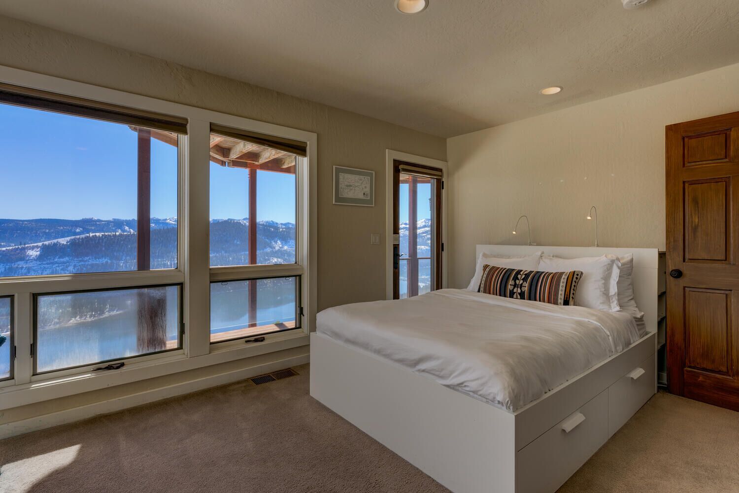 A bedroom with a large bed, two windows with a view of mountains, and a wooden door.