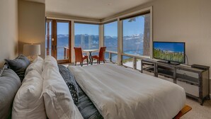 A hotel room with a large bed, a desk with a television, and a view of mountains through large windows. - Big Sky - Lakeview - TLUXP (Truckee)