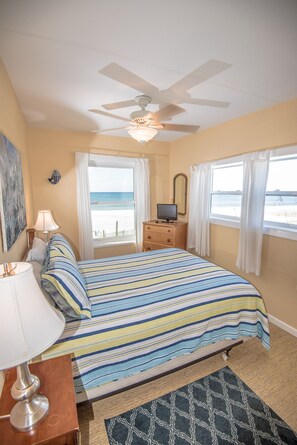A bedroom with a bed, a TV, a ceiling fan, and a view of the beach.