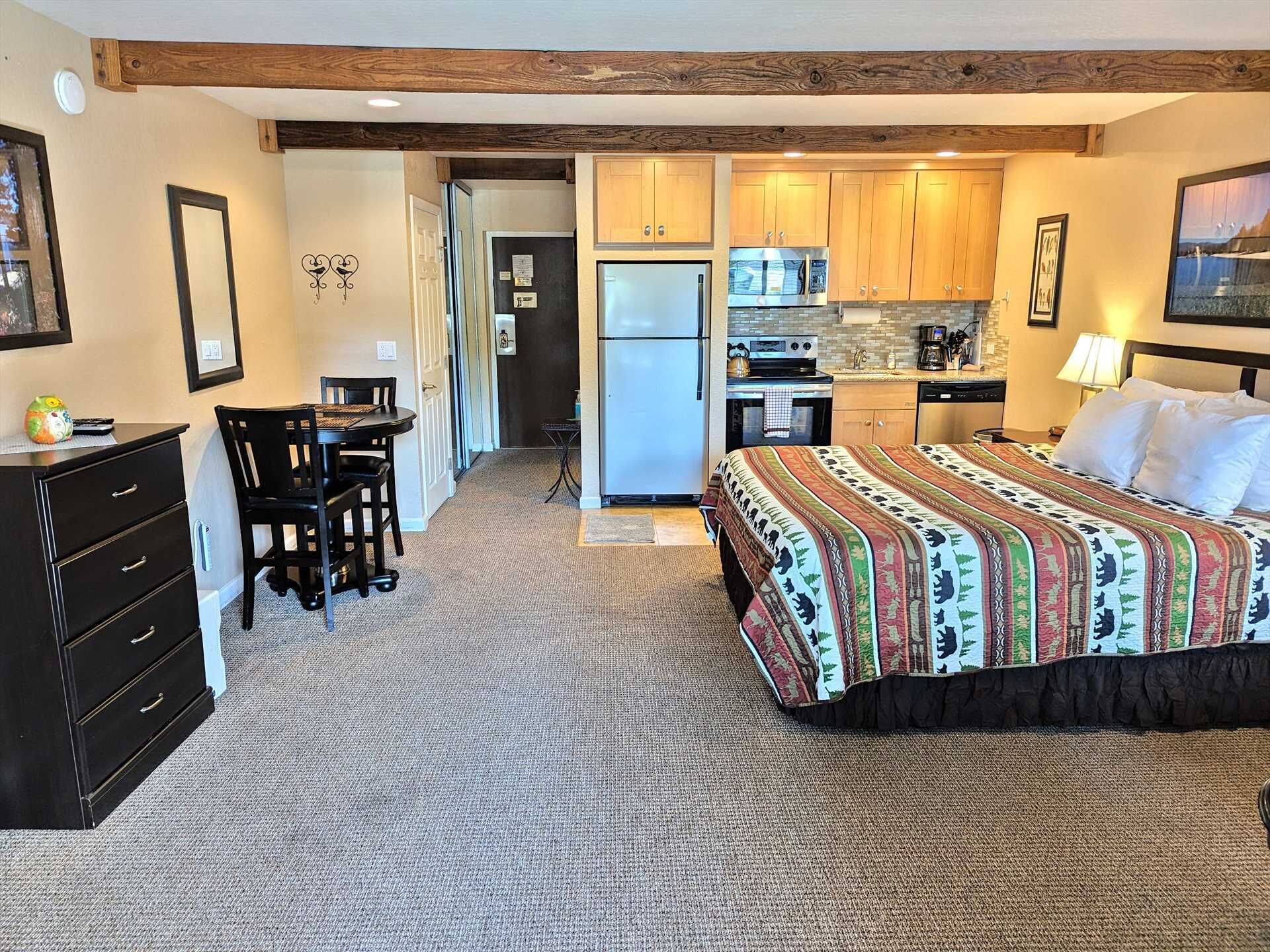 A compact hotel room with a bed, a desk, a chair, a dresser, and a kitchenette.