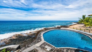 A pool with a curved edge, surrounded by a stone deck and a railing, overlooking the ocean.
