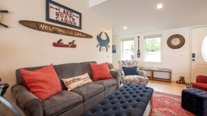 Una sala de estar con un sofá gris, cojines rojos y blancos, un reposapiés azul, un sillón estampado y un banco de madera. Hay decoraciones en la pared, incluyendo un letrero que dice 'Lake Boats Cabins' y un cartel de bienvenida.