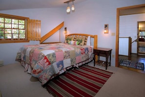 A bedroom with a large bed, a wooden headboard, a window with blinds, a wooden staircase, and a door.