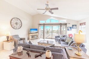 A spacious living room with a large ceiling fan, a clock, and a television. There is a view of the outdoors through large windows.