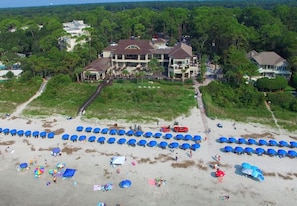 An aerial view of a beach with rows of blue umbrellas and a large building with a driveway leading to it.