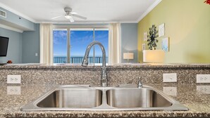A kitchen with a double sink, granite countertop, ceiling fan, and a view of the ocean through a large window.