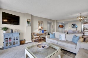 Un salon comprenant un canapé, une table basse, un téléviseur, un bureau avec une lampe et un coin repas avec des chaises.
