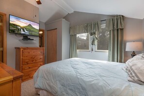 Un dormitorio con cama, cómoda, televisor montado en la pared y ventana con cortinas.