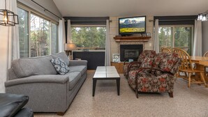A living room with a grey sofa, patterned armchair, and a coffee table. A fireplace with a TV above it is visible, along with large windows offering a view of trees.