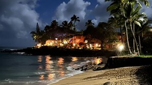 Ein Strand mit Fußspuren, die zu einem Gebäude mit Lichtern, Palmen und Blick auf das Meer führen.