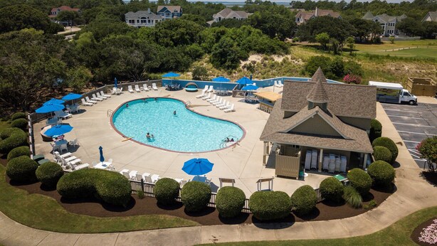 Une piscine entourée de chaises longues et de parasols, bordée de haies bien entretenues et d’un bâtiment doté d’un toit à deux versants.