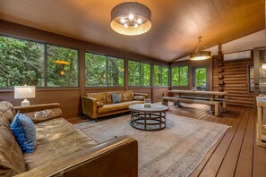 A wooden interior with a leather sofa, round table, and large windows overlooking greenery.