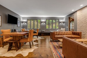A modern living room with a dining area, leather sofas, a wooden dining table, and large windows with a view of greenery.