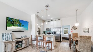 Une cuisine moderne avec des meubles blancs, un îlot central et un coin repas comprenant des chaises en bois et une chaise en osier.